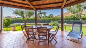 A covered patio with a wooden table and chairs, a lounge chair, and a view of a landscaped garden with trees and a fence. - Villa by the sea with garden (Piana Calzata)