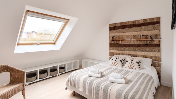 Une chambre avec un plafond en pente, une grande fenêtre, un lit avec une literie à rayures et un fauteuil en osier.