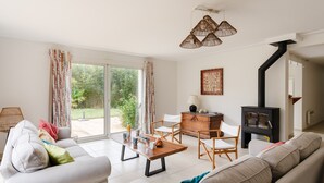 Un salon moderne avec un canapé, une table basse en bois et une table à manger avec des chaises. On y trouve un poêle à bois noir et une grande fenêtre avec des rideaux.