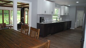 A modern kitchen with white cabinets, a wooden dining table, and a view of a green outdoor area.