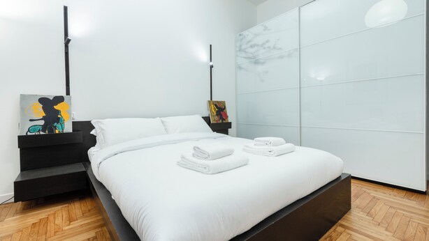 Une chambre moderne avec un lit, du linge de lit blanc, une table de chevet noire et des œuvres d’art.