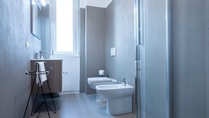 Une salle de bain moderne équipée de toilettes, d’un bidet et d’un lavabo. On y trouve un porte-serviettes avec une serviette, un miroir et une fenêtre qui laisse entrer la lumière naturelle.