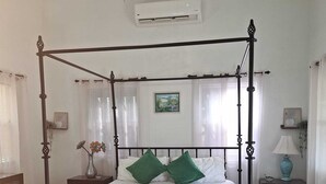 Una cama con dosel, acompañada de un banco, ropa de cama blanca, almohadas verdes, una mesita de noche con lámpara y un cuadro en la pared.