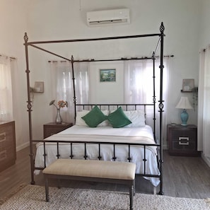 Un lit à baldaquin avec un banc, du linge de lit blanc, des coussins verts, une table de chevet avec une lampe et un tableau au mur.