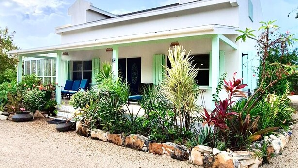 Una casa de un piso con porche cubierto, un pequeño jardín con diversas plantas y un camino de entrada de grava.