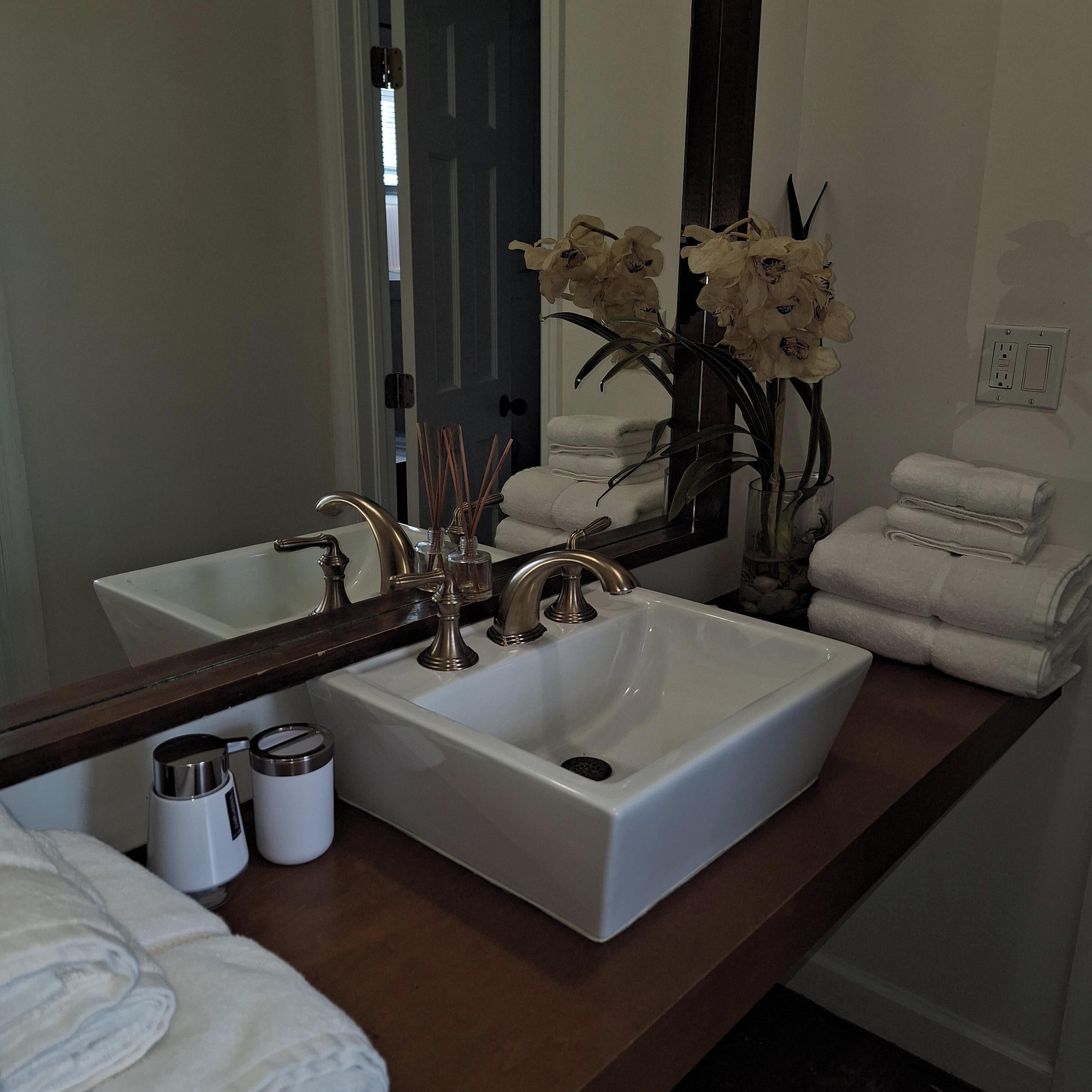 Un bagno con un lavandino bianco, due rubinetti, uno specchio, un vaso con fiori e una pila di asciugamani.