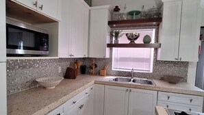 Una cocina con armarios blancos, microondas, fregadero y ventana con persianas.