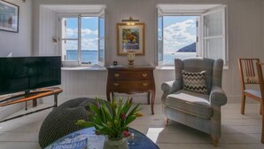 Ein Wohnzimmer mit Fernseher, einer Couch, einem Sessel, einem kleinen Tisch mit Blumenvase und Meerblick durch große Fenster.