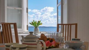 Ein Esstisch mit gestreiften Tassen, einem Teekessel und einer Schüssel mit Essen, vor einem Fenster mit Blick auf das Meer und die Wolken.
