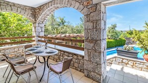 A stone-walled outdoor patio with a round table and chairs, a wooden railing, and a view of a swimming pool. - Villa Epavlis - Three Bedroom Villa, Sleeps 6 (Pilos Nestor)