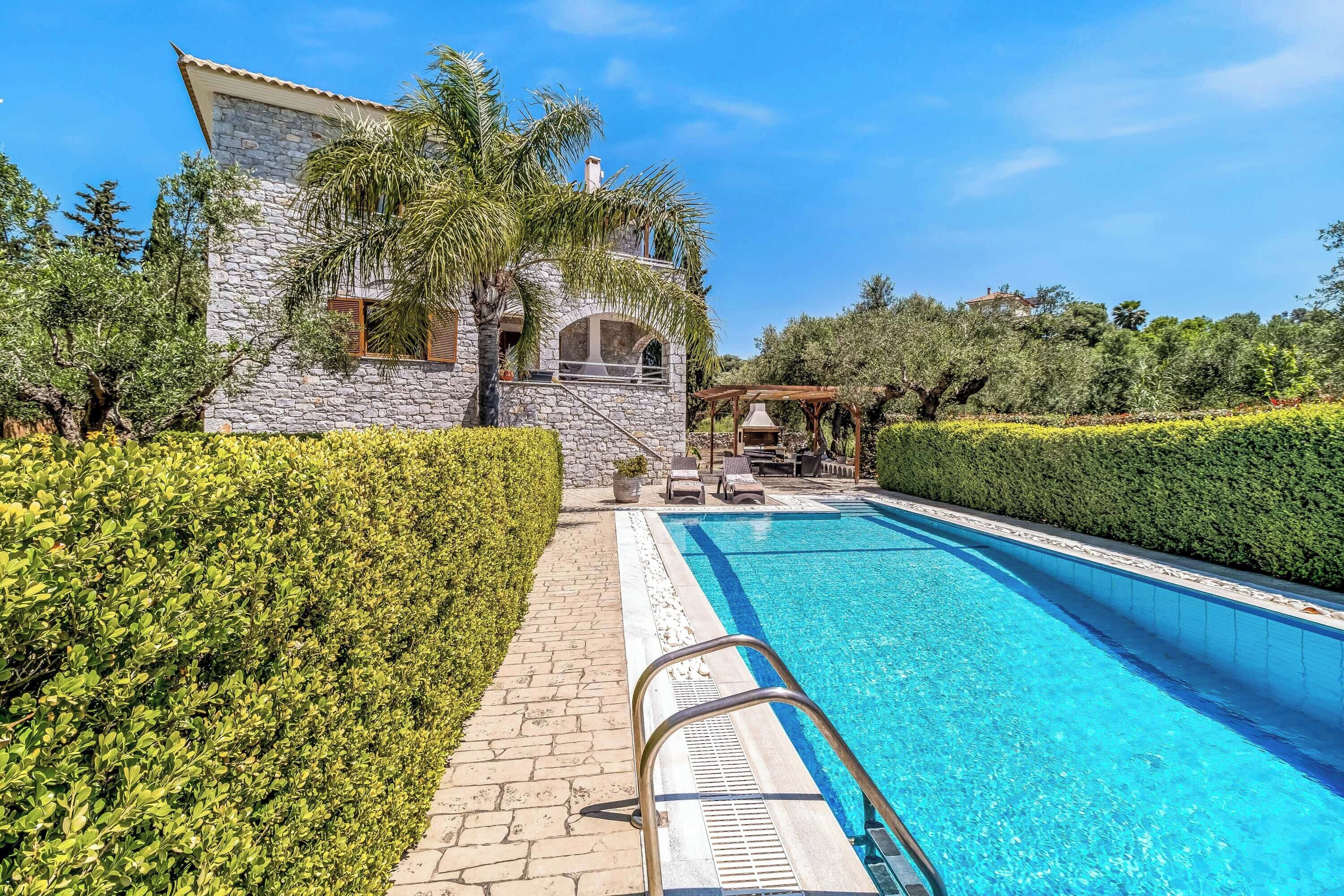 A stone villa with a swimming pool, surrounded by well-maintained hedges and trees.