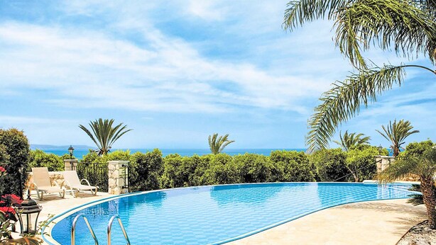 A swimming pool with a curved edge, surrounded by a stone deck and palm trees, with a view of the sea in the background.