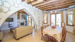 A dining area with a round table and chairs, a sofa, and a television in a stone-walled room with a tiled floor.