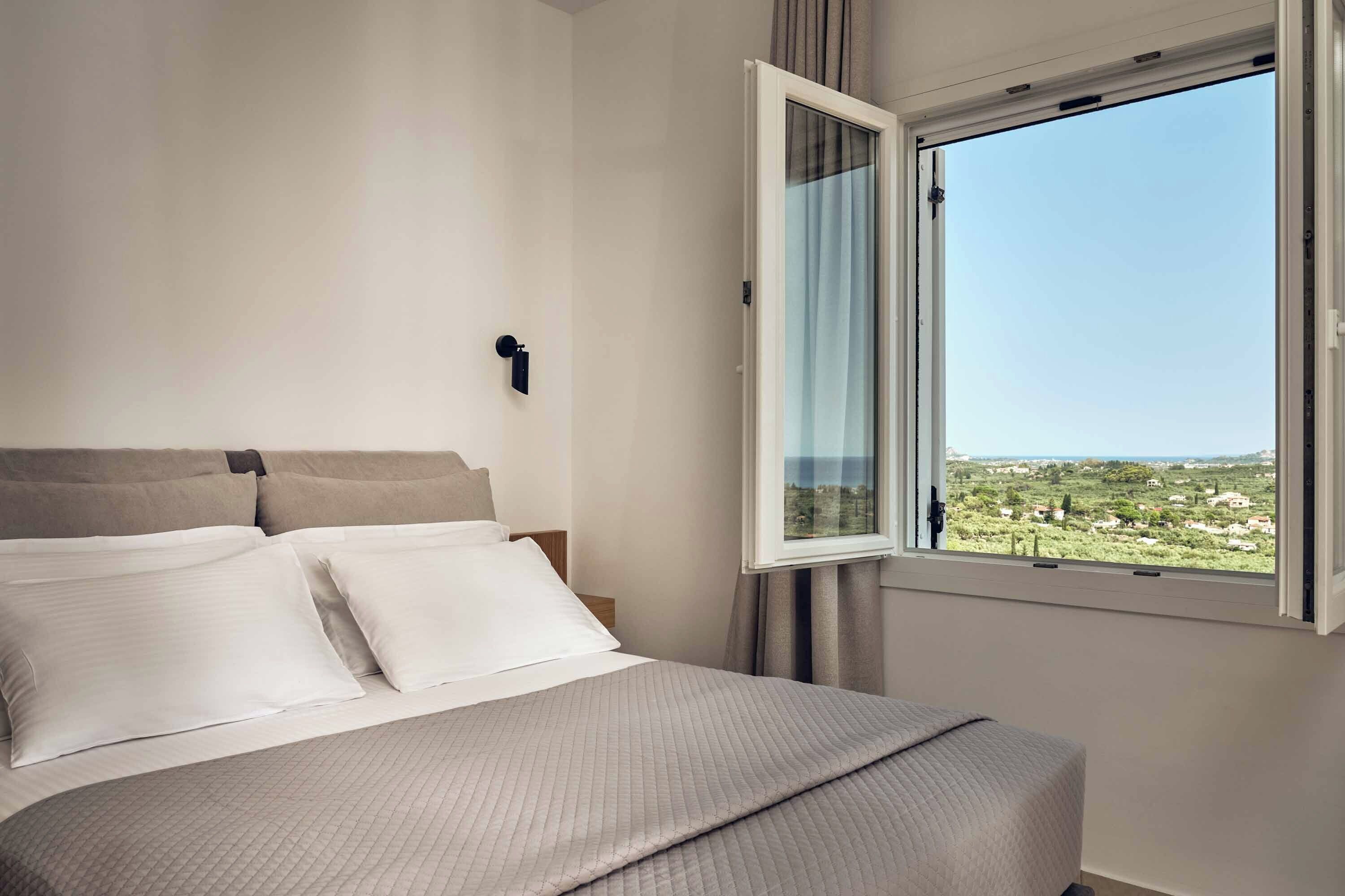 Quarto com cama, mesa de cabeceira, janela com cortinas e vista para uma paisagem.