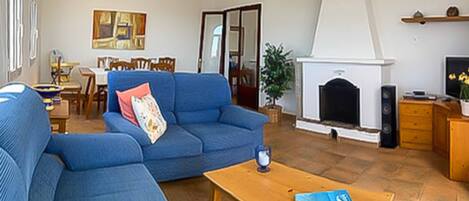 A living room with a blue sofa, a wooden coffee table, a fireplace, and a dining area in the background.