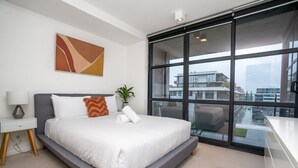 A modern hotel room with a large bed, a desk, a chair, and a view of the city through large windows.