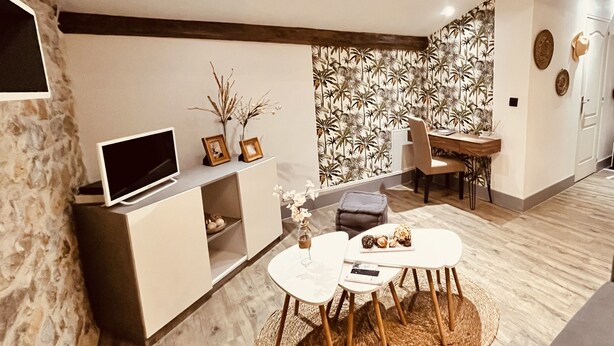 A modern living room with a stone accent wall, a flat-screen TV, a dining area with a small table and chairs, and a patterned wallpaper. - Le Cocon Charmant Studio (Nîmes)
