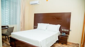 Una cama bien tendida con sábanas blancas en una habitación con suelo de madera, un escritorio y un teléfono en la mesita de noche.