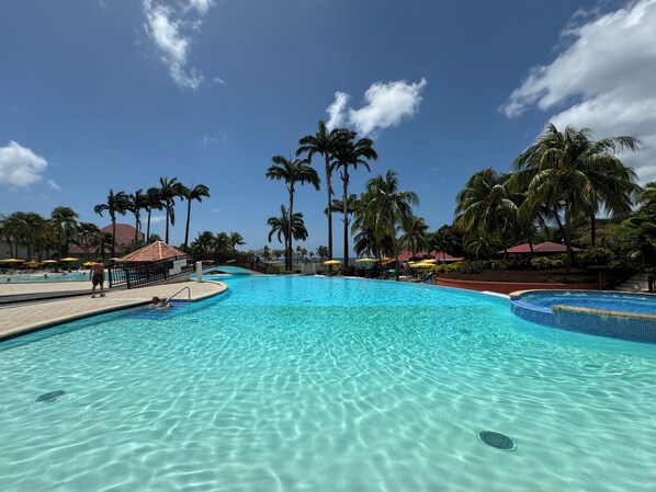 A large outdoor swimming pool surrounded by palm trees and umbrellas, with clear blue water and a sunny sky.