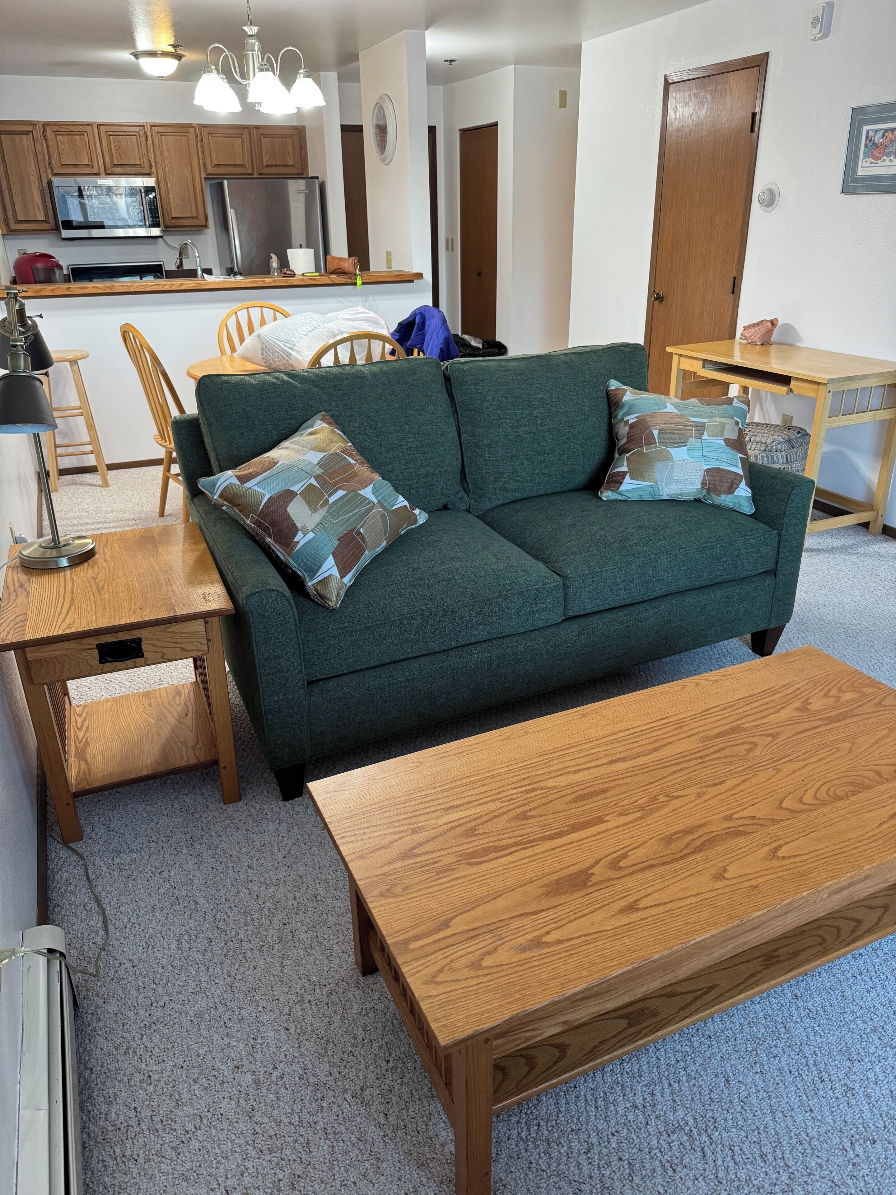 A living room with a green sofa, wooden coffee table, and a kitchen area with wooden cabinets and a microwave.