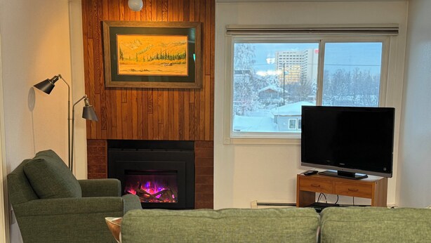 A living room with a fireplace, a green sofa, a television on a wooden stand, and a framed picture above the fireplace. -  1 bdrm condo. Indoor parking garage. Secure bldg access.  90 day minimum stay (Anchorage)