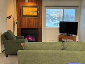 A living room with a fireplace, a green sofa, a television on a wooden stand, and a framed picture above the fireplace. -  1 bdrm condo. Indoor parking garage. Secure bldg access.  90 day minimum stay (Anchorage)