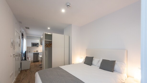 Una habitación de hotel moderna con una cama grande, mesitas de noche, un armario y una kitchenette al fondo.
