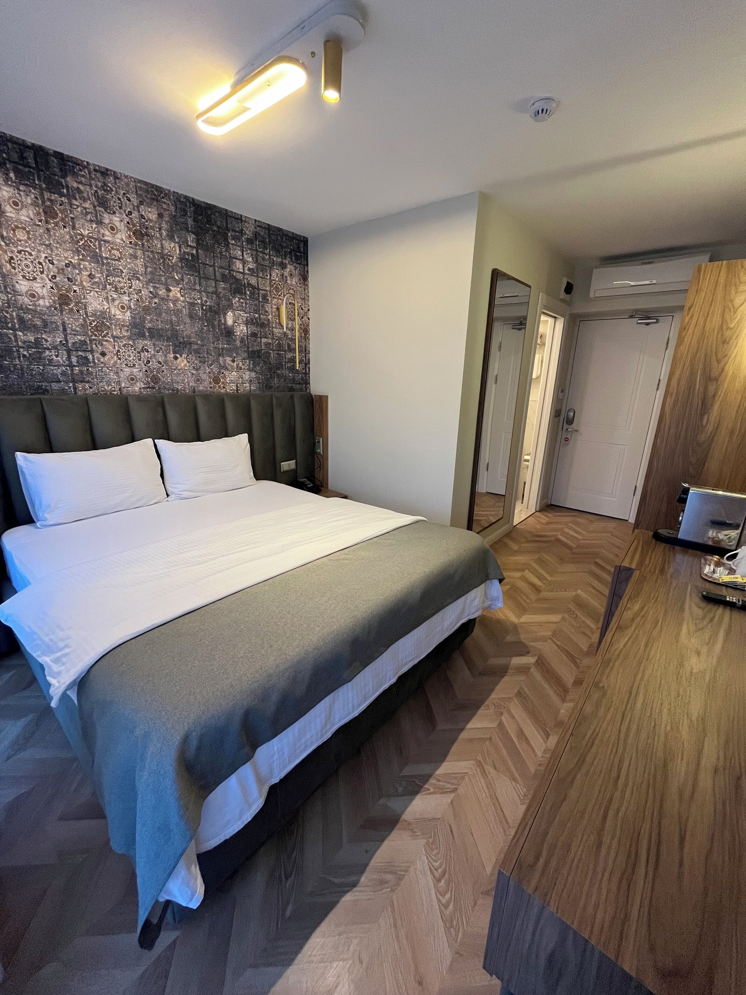 A modern hotel room with a large bed, a wooden floor, a wall with a textured pattern, and a door leading to another room.