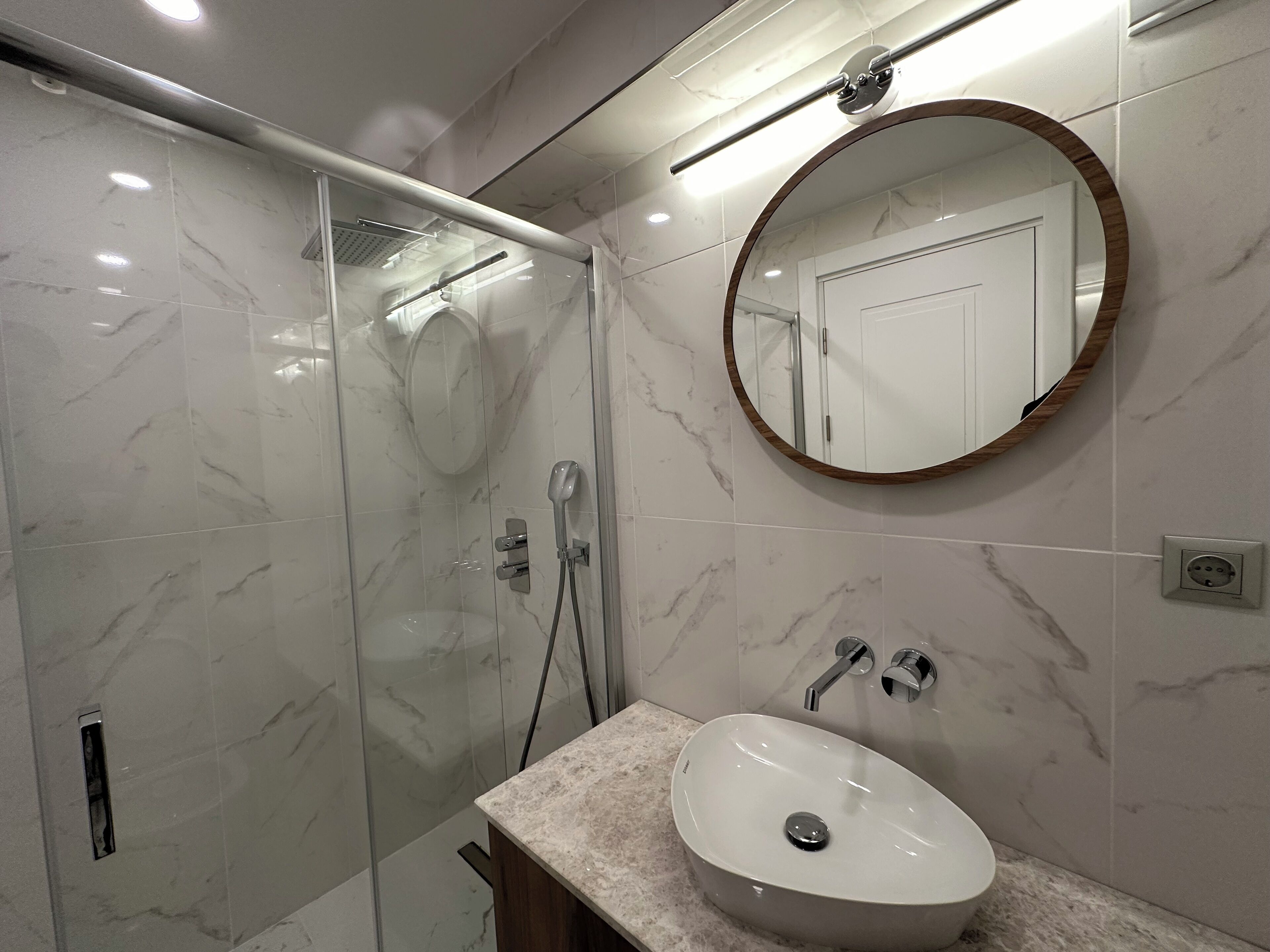 A modern bathroom with a glass-enclosed shower, a round mirror, and a marble-tiled sink.