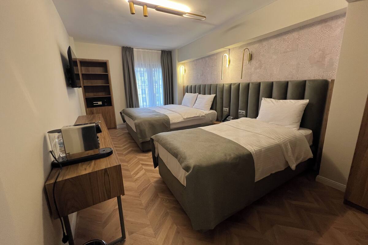 Habitación de hotel con dos camas, un escritorio con cafetera, un televisor montado en la pared y un papel tapiz decorado.