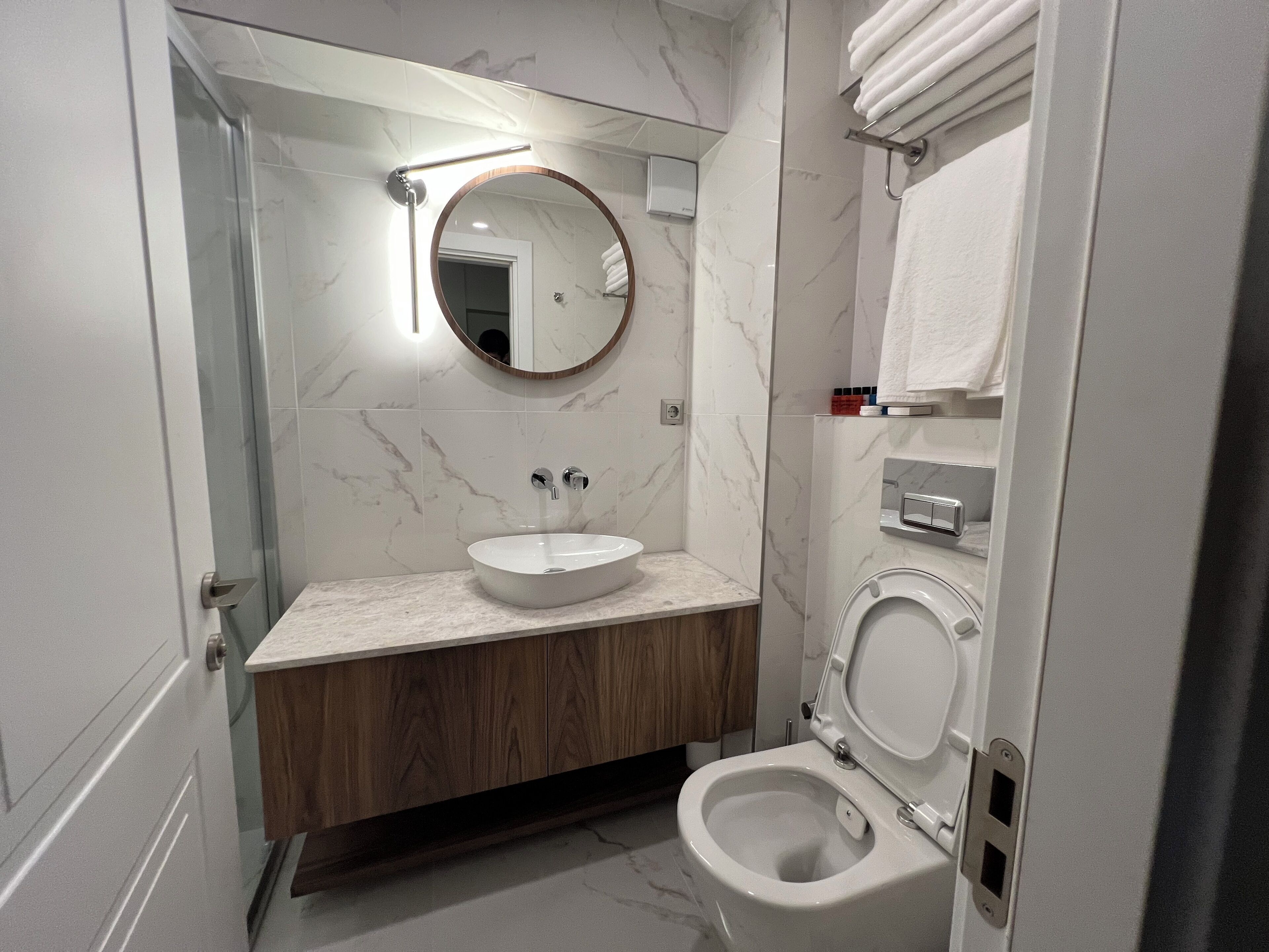 A modern bathroom with a marble sink, wooden vanity, a round mirror, and a toilet.