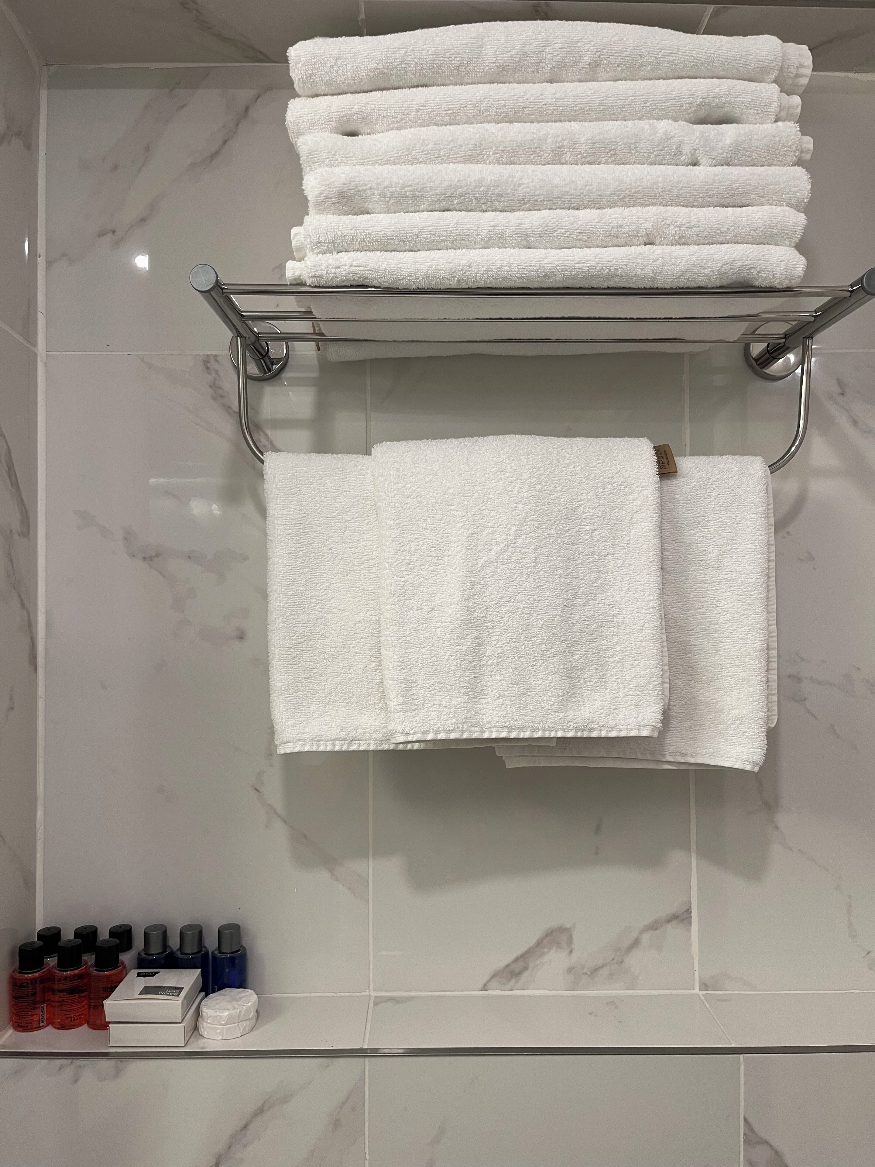 Un baño con una pila de toallas blancas en un estante metálico, una toalla doblada colgada en un perchero y artículos de tocador en una repisa.