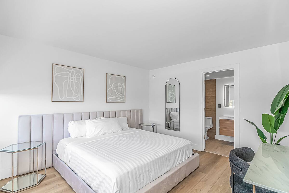 A modern bedroom with a large bed, bedside tables, a mirror, and a view into the bathroom.
