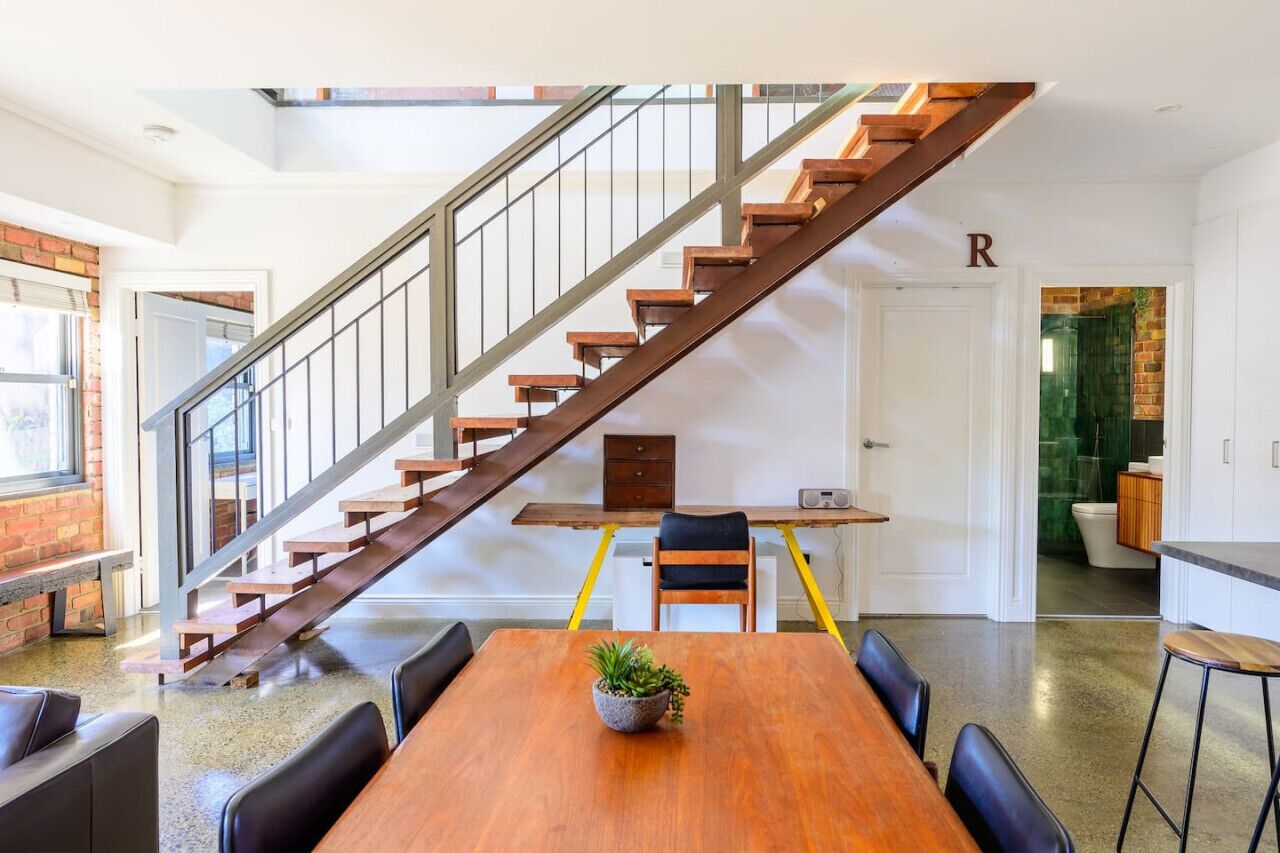 A modern interior with a wooden staircase, a dining area with a table and chairs, and a kitchen visible through an open door.