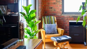 A cozy room with a brick wall, a wooden chair with a cushion, a black leather ottoman, and a desk with a computer.