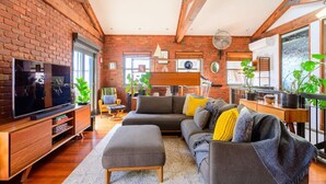 A cozy living room with a brick wall, wooden beams, a large TV, a sofa, and potted plants.