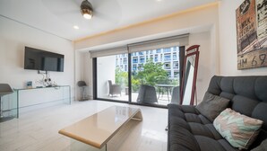 A modern living room with a glass coffee table, a sofa with a cushion, a mounted TV, and a large window offering a view of a building and greenery. - Yucatán - 1 Bedroom in Playa Near 5th At 38th Street (Playa del Carmen)
