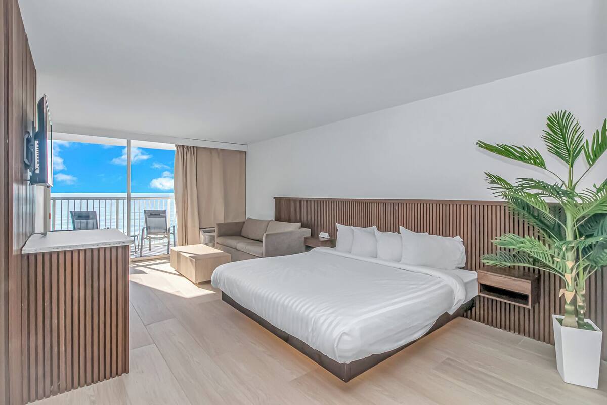 A modern hotel room with a large bed, a seating area with a sofa and chairs, a small table, a potted plant, and a balcony with a view of the sea.