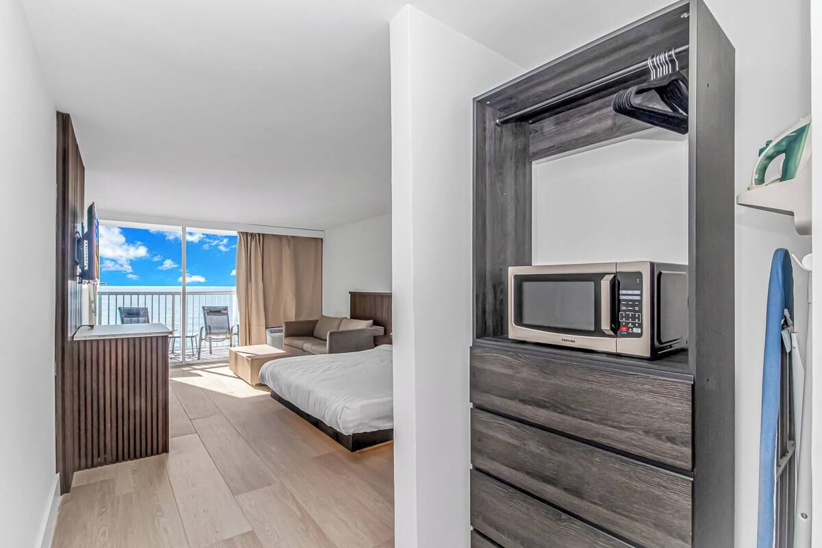 A hotel room with a bed, a sofa, a TV, and a microwave. There is a balcony with a view of the sky.
