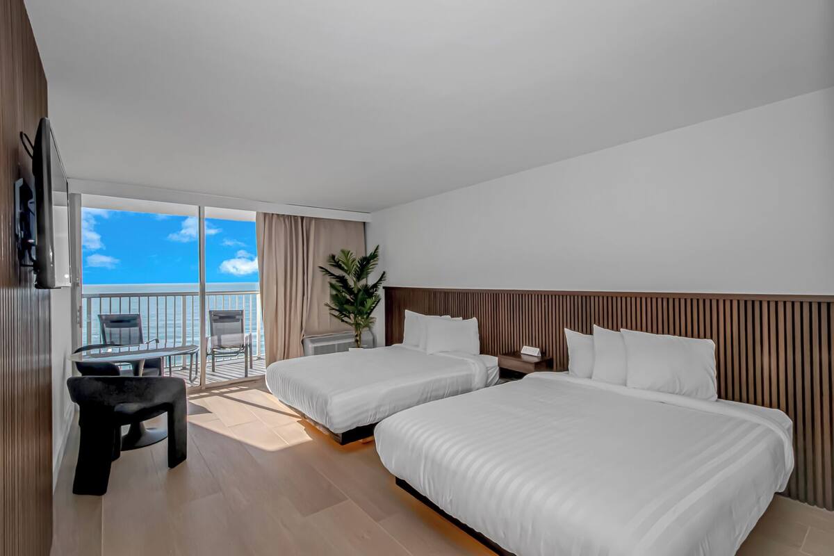 A hotel room with two beds, a desk, and a balcony with a view of the ocean.