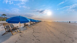 A beach with rows of blue umbrellas and beach chairs, a clear sky, and the ocean in the background. - 321 Oceanfront Villa | Walk to Coligny + Pools | Washer/Dryer (Hilton Head Island)