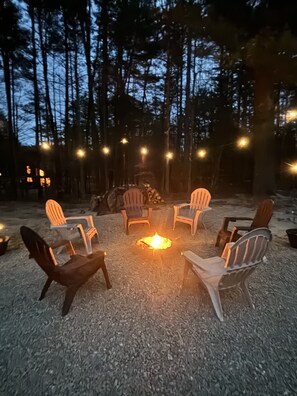 En eldstad omgiven av fyra Adirondack-stolar, placerad på ett grusunderlag med träd i bakgrunden.