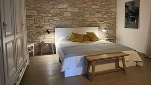 Un dormitorio con pared de piedra, cama con ropa blanca y almohadas doradas, mesita de noche con lámpara y un banco de madera.