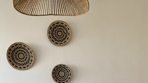 Ein Essbereich mit einem weißen Tisch, vier weißen Stühlen, einer gewebten Pendelleuchte und drei Wanddekorationen aus Bändern.