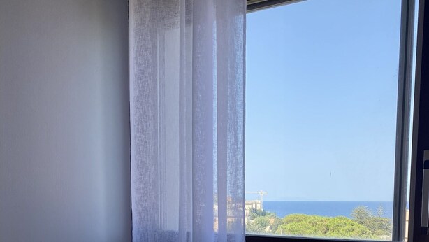 Ein Schlafzimmer mit Bett, einem Fenster mit transparenten Vorhängen und Blick auf das Meer sowie entfernte Gebäude.