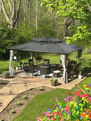 A covered outdoor patio with seating and tables, surrounded by greenery and trees.