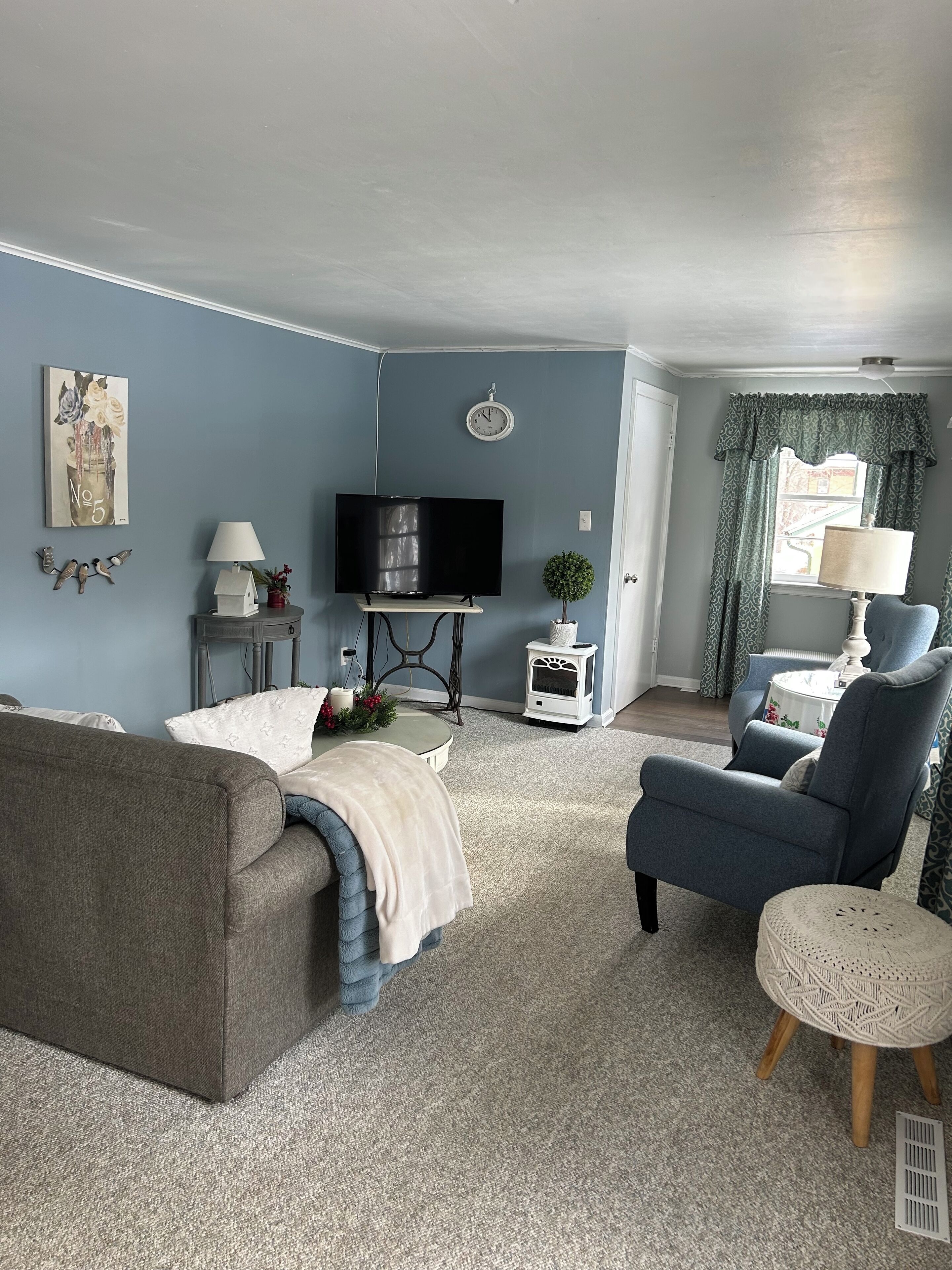 A living room with a grey sofa, a blue armchair, a television on a stand, a small table with a lamp, and a decorative wall piece.