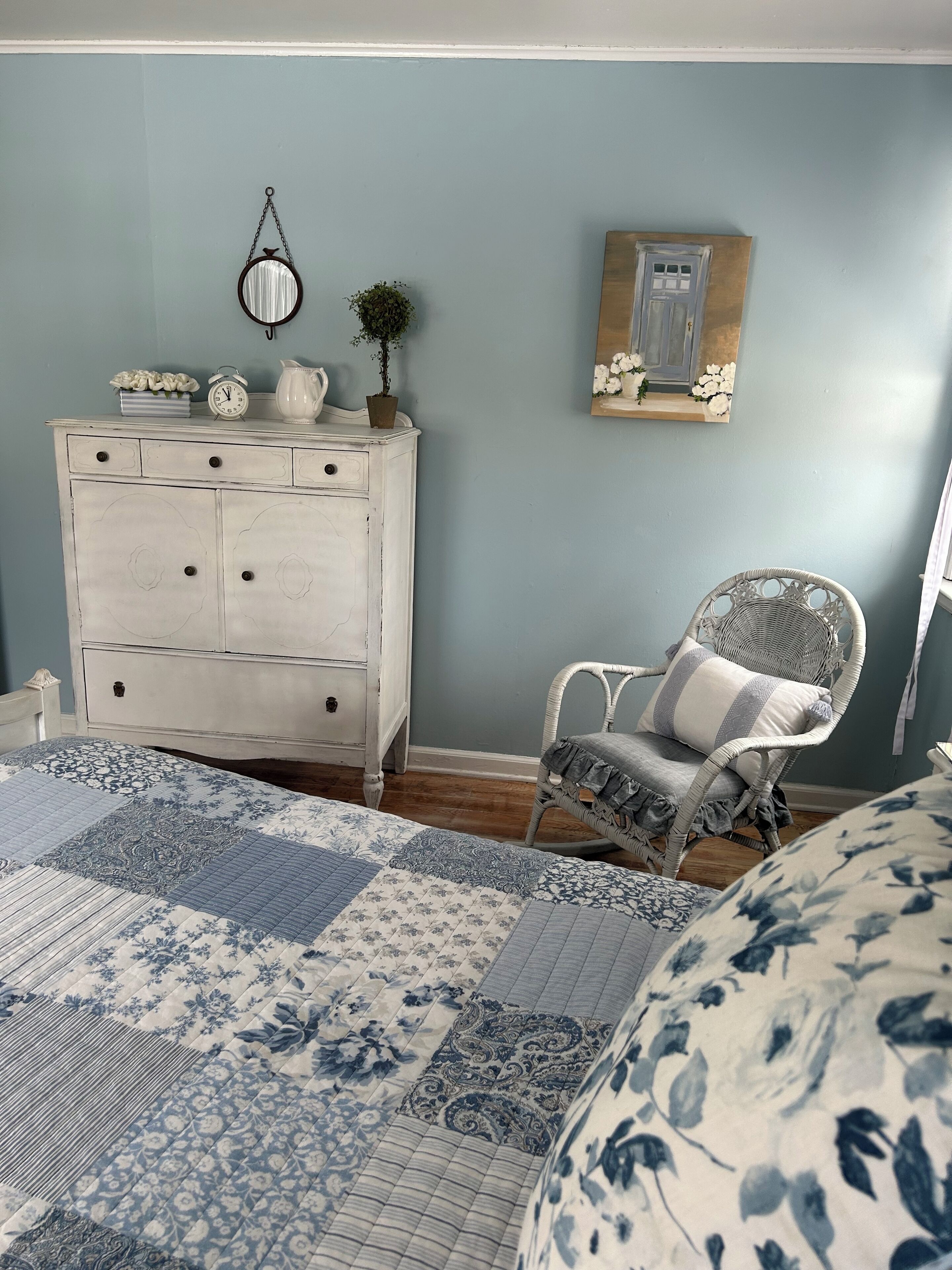 A bedroom with a white dresser, a wicker chair, a floral bedspread, and a wall-mounted shelf with a picture.