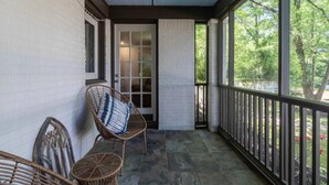 A covered porch with wicker chairs, a door, and a view of greenery.