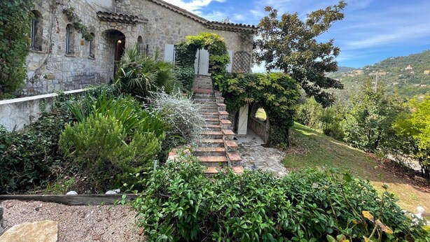 Un bâtiment en pierre avec un sentier de jardin menant à une porte, entouré d’une végétation luxuriante et sous un ciel dégagé.